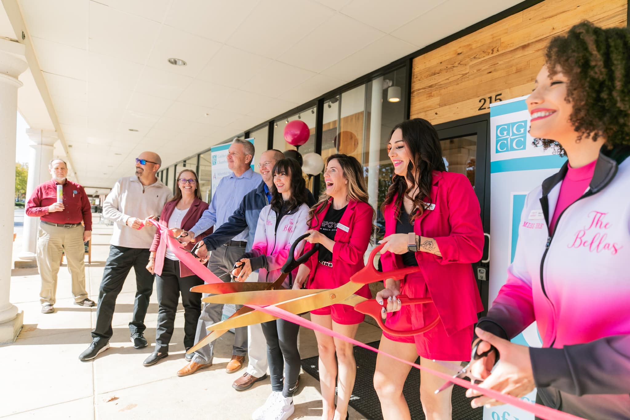 The Bellas team cutting a pink ribbon with oversized gold scissors at the Empower studio grand opening, alongside the Gadsden Chamber of Commerce
